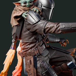 May include: A detailed figurine of The Mandalorian with Grogu. The Mandalorian is in dark armor with a helmet, and Grogu is perched on his shoulder, wrapped in a brown cloak. A flame effect emanates from the jetpack.