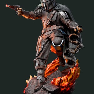 May include: A detailed statue of a character from The Mandalorian. The figure is clad in dark armor, holding a blaster pistol with a red barrel. The statue stands on a rocky base with fiery orange and yellow accents.