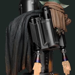 May include: A detailed statue of The Mandalorian with Grogu. The Mandalorian is clad in black armor with a cape, jetpack, and ignited thrusters. Grogu is wrapped in a brown blanket, perched on the Mandalorian's shoulder. The statue has a dark green background.