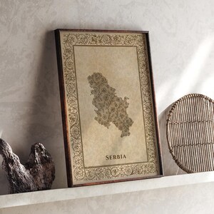 Puede incluir: Un cuadro rectangular enmarcado que presenta un mapa de Serbia relleno con intrincados patrones florales. El mapa está centrado sobre un fondo beige estilo pergamino, rodeado por un borde decorativo de enredaderas y flores. Se lee la palabra SERBIA.