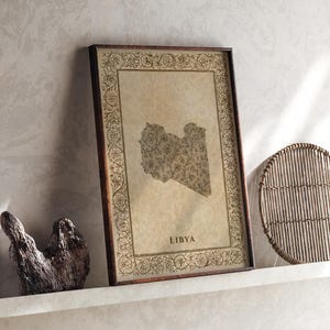 Pode incluir: Uma impressão retangular emoldurada apresentando um mapa da Líbia no centro, preenchido com um padrão floral detalhado. O mapa é cercado por uma borda decorativa de vinhas e flores intrincadas, com a palavra LIBYA impressa abaixo do mapa.