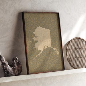 Puede incluir: Una pieza de arte mural rectangular enmarcada que presenta una silueta beige del mapa de Alaska centrada sobre un fondo verde oliva oscuro con un patrón repetitivo de pequeños motivos florales de color amarillo dorado.