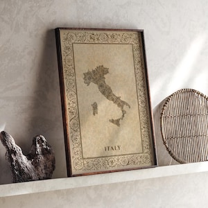 Può includere: Una stampa rettangolare incorniciata che mostra una mappa dell'Italia in stile vintage su uno sfondo di pergamena beige. Il bordo è decorato con intricati motivi floreali. La parola ITALY è stampata in lettere maiuscole nere sotto la mappa.