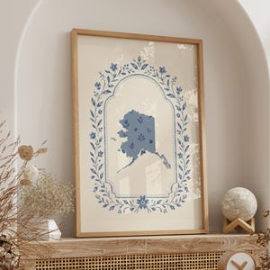 Puede incluir: Una impresión de arte mural rectangular en un marco de madera clara que presenta una silueta azul del estado de Alaska centrada dentro de una corona floral azul ornamentada y un borde decorativo sobre un fondo de color crema.