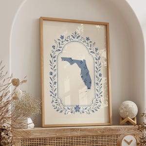 Pode incluir: Uma impressão de arte de parede vertical emoldurada apresentando uma silhueta azul do estado da Flórida centralizada dentro de uma borda ornamental em forma de coroa floral oval. O design é em tons de azul sobre um fundo creme claro.