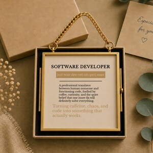 K&ouml;nnte beinhalten: Ein quadratischer goldfarbener Metallrahmen mit einer Aufh&auml;ngungskette, der eine bedruckte Karte mit der Definition eines Softwareentwicklers enth&auml;lt. Der Text lautet: SOFTWARE DEVELOPER, a professional translator between human nonsense.