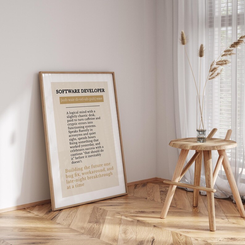 May include: A framed print with the words "SOFTWARE DEVELOPER" at the top. The text below describes the role of a software developer. The print is in a light beige frame, leaning against a wall next to a small wooden table.