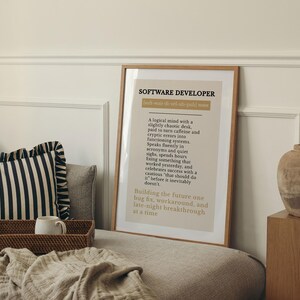 May include: A framed print with the text "SOFTWARE DEVELOPER" in a light brown frame. The print has a beige background with black text describing a software developer's traits. A decorative vase sits on a wooden block.