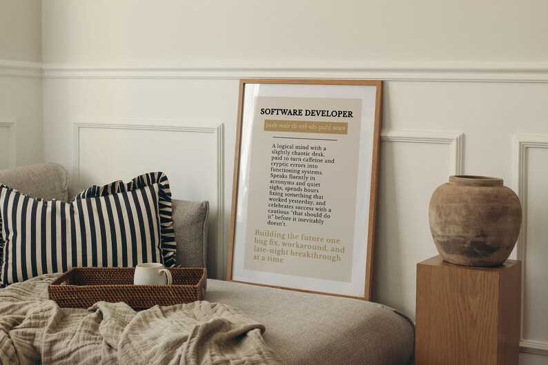 May include: A framed print with the text "SOFTWARE DEVELOPER" in a light brown frame. The print has a beige background with black text describing a software developer's traits. A decorative vase sits on a wooden block.