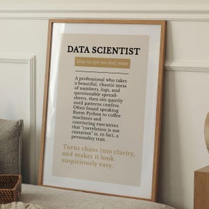 May include: A framed print with the words "DATA SCIENTIST" in bold black letters at the top. The text below describes the role of a data scientist, with a definition and humorous observations. The frame is a light brown color.