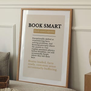 Op de afbeelding: Een ingelijste kunstprint met de tekst BOOK SMART, gedefinieerd als een bijvoeglijk naamwoord. De print bevat een woordenboekachtige beschrijving over het onthouden van feiten en oplossen van problemen, op een lichtbeige achtergrond.