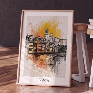 May include: A framed art print featuring a watercolour and ink illustration of the city of Girona, Spain. The artwork depicts buildings and a bridge over a river with yellow and orange paint splatters. The text Girona Spain is printed below.