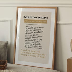 May include: A framed wall art print featuring a dictionary-style definition of the Empire State Building. The text is printed in black and gold on a beige background, presented in a light wood frame with a clean, rectangular design.