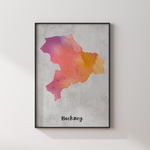 May include: A framed wall art print featuring a watercolor map of Hackney in shades of pink, orange, and yellow. The map is set against a textured light gray background, with the word Hackney printed in black script lettering at the bottom.