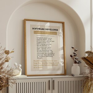 May include: A framed print with the text "SOFTWARE DEVELOPER" in a wooden frame. The print has a beige background with black text describing the role. The print is displayed on a white cabinet with decorative items.