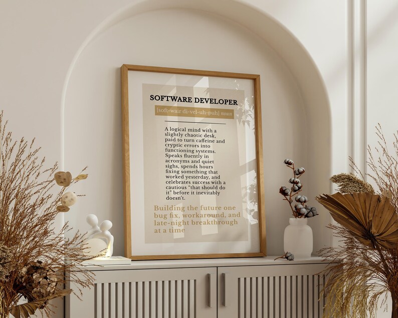 May include: A framed print with the text "SOFTWARE DEVELOPER" in a wooden frame. The print has a beige background with black text describing the role. The print is displayed on a white cabinet with decorative items.