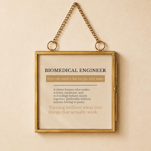 Op de afbeelding: Een vierkante wandhanger in een messing lijst met een ketting, voorzien van de tekst: BIOMEDICAL ENGINEER [bye-oh-med-i-kal en-jin-eer] noun. A clever human who makes science, medicine, and technology behave nicely together. Turning brilliant ideas into things that actually work.