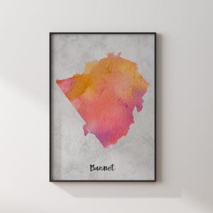 May include: A framed wall art print featuring a watercolor-style map shape in shades of orange, pink, and yellow against a textured light gray background. The word Bannet is printed in black, handwritten-style lettering at the bottom center.