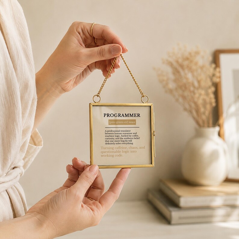 May include: A square, gold-toned metal hanging frame containing a printed definition of the word PROGRAMMER. The text reads: PROGRAMMER [pro-gram-er] noun. A professional translator between human nonsense and machine logic, fuelled by coffee, curiosity, and the stubborn belief that one more bug fix will definitely solve everything. Turning caffeine, chaos, and questionable logic into working code.