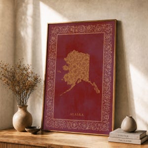May include: A rectangular framed wall art print featuring a deep red background with a gold floral patterned silhouette of the state of Alaska in the centre. The border is decorated with intricate gold floral scrollwork and the text ALASKA.