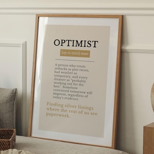 May include: A framed wall art print featuring the word OPTIMIST in bold black letters, followed by its phonetic spelling and a dictionary-style definition describing a person who views setbacks as plot twists and finds silver linings.