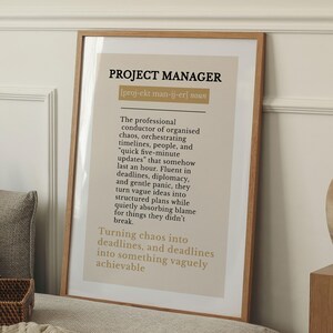 以下が含まれることがあります： 太字の「PROJECT MANAGER」というテキストが入った額入りのプリント。プロジェクトマネージャーの定義はタイトルの下にあります。フレームは明るい茶色です。