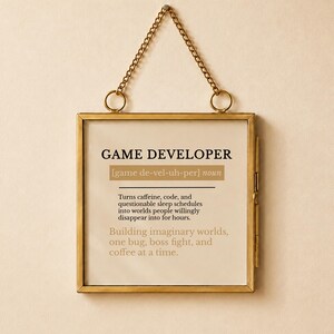 Pode incluir: Um pendente de parede quadrado numa moldura de metal dourado com uma corrente para pendurar. A impressão apresenta o texto GAME DEVELOPER, uma pronúncia fonética e uma definição que descreve o papel como transformar cafeína e código em mundos.