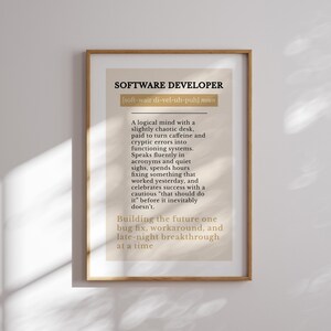 May include: A framed print featuring the text "SOFTWARE DEVELOPER" and a definition. The definition describes a software developer's traits, including a logical mind, caffeine use, and late-night work. The frame is a light brown colour.