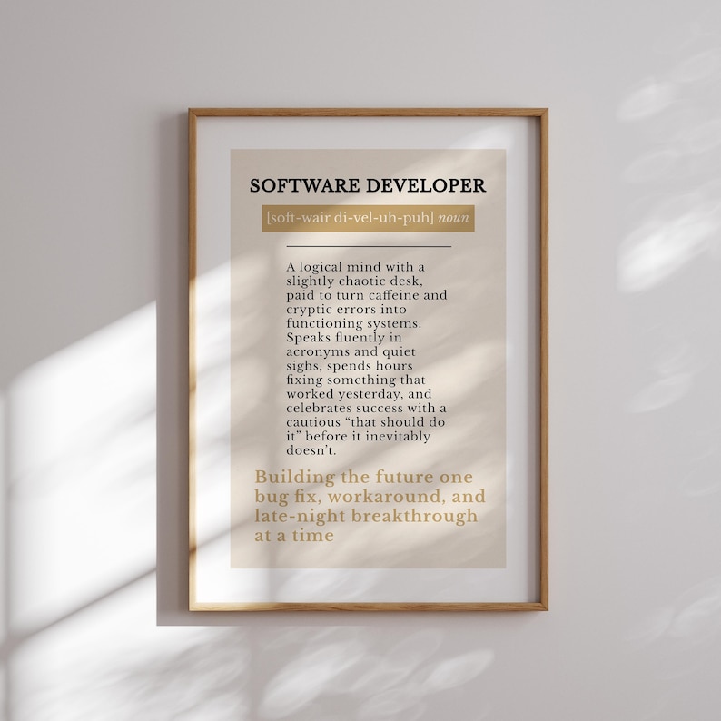 May include: A framed print featuring the text "SOFTWARE DEVELOPER" and a definition. The definition describes a software developer's traits, including a logical mind, caffeine use, and late-night work. The frame is a light brown colour.