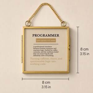 May include: A square gold-toned metal frame with a hanging chain, displaying a definition of the word PROGRAMMER. The text reads: A professional translator between human nonsense and machine logic, fuelled by coffee, curiosity, and the stubborn belief that one more bug fix will definitely solve everything. Turning caffeine, chaos, and questionable logic into working code. The frame measures 8 cm by 8 cm.
