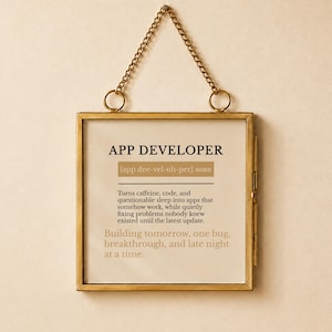 Puede incluir: Un colgante de pared cuadrado en un marco de metal dorado fino con una cadena a juego. El estampado presenta el texto APP DEVELOPER, una pronunciación fonética y una definición humorística sobre convertir cafeína en software.