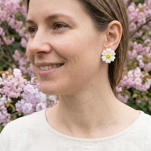 Könnte beinhalten: Eine Person trägt weiße Ohrringe in Form von Gänseblümchen mit gelben Zentren. Die Ohrringe sind vor einem Hintergrund aus rosa Blüten angebracht, und die Person trägt ein helles Oberteil.