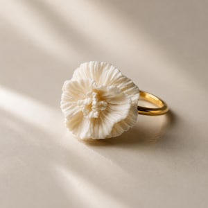 Peut inclure: Bague fleur blanche avec un anneau doré. La fleur présente des pétales détaillés et une surface texturée. La bague est posée sur un fond neutre, soulignant le design délicat. Un bijou unique.