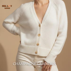 May include: A cream-colored knit cardigan featuring a V-neckline, long sleeves, and four light brown buttons down the front. The text "OPAL LOOP DESIGNS" appears in the upper left corner, and "CHAMPAGNE CARDIGAN" is printed at the bottom of the image.