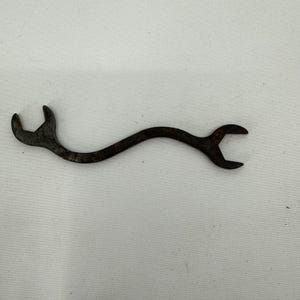 May include: A weathered, curved wrench with a dark, rusty appearance. The wrench features two open ends, each with a different sized opening. The tool is made of metal and shows signs of wear, indicating it's an antique or a used item.
