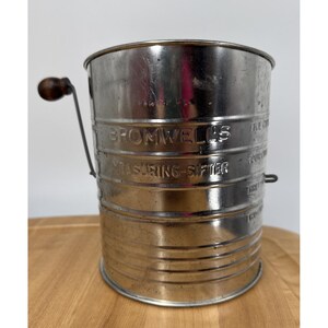 May include: A cylindrical metal Bromwell's measuring sifter with a dark brown wooden handle on a side crank. The exterior features embossed text reading BROMWELL'S MEASURING-SIFTER and measurement markings for one, two, three, and five cups.