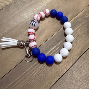 Op de afbeelding: Een polsbandje sleutelhanger met witte, blauwe en honkbal-thema kralen. De honkbal kralen hebben rode stiksels en een witte achtergrond. De polsband bevat een witte kwast en een zilverkleurige sluiting.