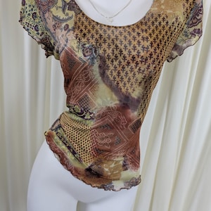 Könnte beinhalten: Ein transparentes Oberteil mit kurzen Ärmeln und Rundhalsausschnitt, mit einem braunen, gelben und lila Paisley-Muster. Das Oberteil hat eine taillierte Silhouette und einen gerüschten Saum. Eine silberne Halskette mit kleinem Anhänger ist sichtbar.