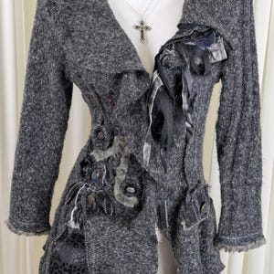 Puede incluir: Una chaqueta gris texturizada con adornos decorativos. La chaqueta presenta un cuello drapeado, mangas largas y varios acentos de tela y cinta. Un colgante de cruz plateada cuelga de un collar. La chaqueta tiene un diseño artístico único.