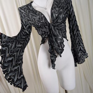May include: A black and grey long-sleeve top featuring a wavy, chevron-like knit pattern throughout. The garment has a deep V-neckline, a front tie closure, and dramatic, flared bell sleeves with ruffled edges, displayed on a white mannequin.