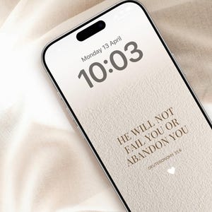 Op de afbeelding: Een smartphonescherm toont de tijd 10:03 en de datum maandag 13 april. Het scherm toont de tekst "HE WILL NOT FAIL YOU OR ABANDON YOU" en "DEUTERONOMY 31:6" in een lichtbruin lettertype, met een hartpictogram.