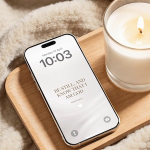 May include: A smartphone with a white screen displaying the time, date, and the text "BE STILL, AND KNOW THAT I AM GOD" on a wooden tray. A lit candle in a glass jar sits beside the phone. The background is a soft, cream-colored blanket.