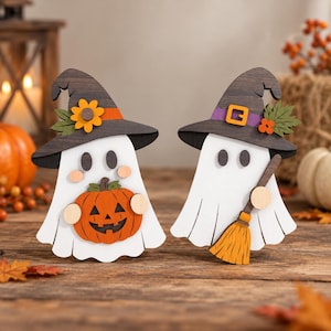 Op de afbeelding: Twee houten spookdecoraties, elk met een donkerbruine heksenhoed. Eén spook houdt een oranje pompoen vast en heeft een gele bloem op de hoed. Het andere spook houdt een bruine bezem vast en heeft een paarse hoedband met gesp.