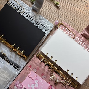 以下が含まれることがあります： HIGH PRIORITYと書かれた黒いページを持つグレーのバインダーと、LOW PRIORITYと書かれた白いページを持つピンクのバインダー。どちらも金色のリング金具、装飾的なチャーム、様々な紙のインサートが入った内ポケットを備えた2冊の開いたリングバインダー。