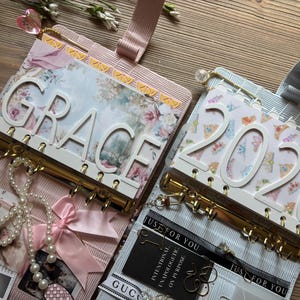 May include: Two personal planners with gold-tone ring binders. The left planner features a floral cover with the white text GRACE and a pink ribbon. The right planner has a patterned cover with the white text 2026 and a black label reading JUST FOR YOU.