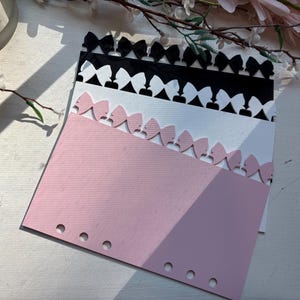 May include: A stack of three rectangular paper planner inserts with a die-cut bow border along the top edge. The top insert is black, the middle is white, and the bottom is light pink. Each insert features a row of circular holes along the bottom.