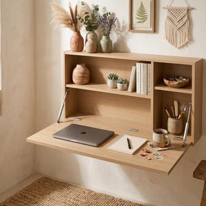 Peut inclure: Un bureau mural en bois clair doté d'une surface de travail rabattable soutenue par des charnières métalliques. L'unité comprend plusieurs étagères de rangement ouvertes contenant des livres, des petites plantes succulentes et un pot à crayons.