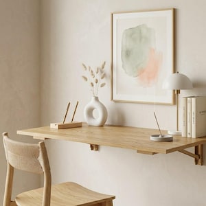 Könnte beinhalten: Ein minimalistisches Home-Office-Setup mit einem hellen Holzschreibtisch, einem Stuhl mit Flechtwerk und einem gerahmten Aquarelldruck. Eine weiße Vase mit Trockenblumen, eine Schreibtischlampe und Bücher ergänzen die Einrichtung.