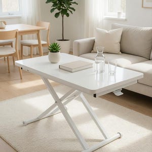 Puede incluir: Una mesa de centro blanca regulable en altura con acabado brillante. La mesa tiene una base en forma de tijera y está situada en un salón con una combinación de colores neutros. Una pequeña suculenta en una maceta blanca, un libro, una jarra de cristal y un vaso están sobre la mesa.