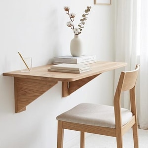 May include: A light wood, wall-mounted desk with a triangular support bracket. A vase with cotton stems and books sit on the desk. A wooden chair with a beige seat is in front of the desk. The desk is approximately 30 inches wide.